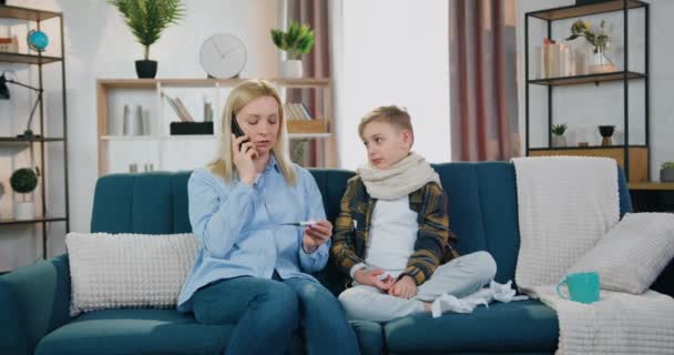 Nice wyczerpany nastolatek chory chłopiec na grypie siedzi na kanapie w pobliżu jego zmartwiony kochający matka, która wzywa lekarza rodzinnego, gdy zobaczyć wskaźniki temperatury na termometrze — Wideo stockowe