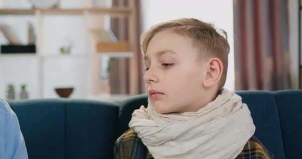 Domácí péče o děti koncept, kde roztomilý unavený rozrušený nemocný chlapec s bolestí v krku sedí na gauči, když matka kontroluje jeho teplotu, dávat mu ruku na čelo — Stock video