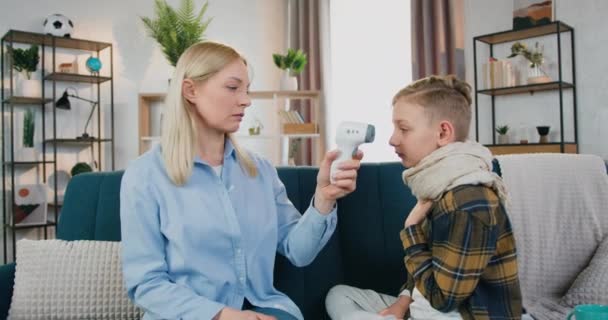 Çok endişeli bir anne temas olmayan kızılötesi termometreyle ısıyı kontrol ediyor hasta oğulların alnına yerleştiriyor ve göstergelerden büyük gözler yapıyor. — Stok video