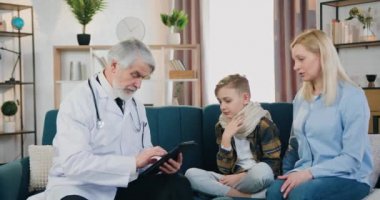 Çekici, saygın, sakallı bir doktorun, çocuk hasta kartını tabletle doldurduğu sağlık hizmeti konsepti, erkek çocukların anneleri hastalık belirtileri hakkında konuşuyorlar.