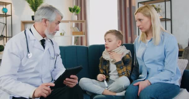 Красивая заботливая любящая блондинка сидит рядом со своим больным усталым сыном, который разговаривает с уважаемым высококвалифицированным бородатым врачом, когда он посещает их дома, медицина и концепция здравоохранения — стоковое видео