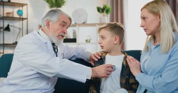 Médecin visite à domicile concept où beau expérimenté respecté pédiatre barbu vérifier garçons battements de coeur avec stéthoscope qui est assis près de soins mère inquiète sur le canapé — Video