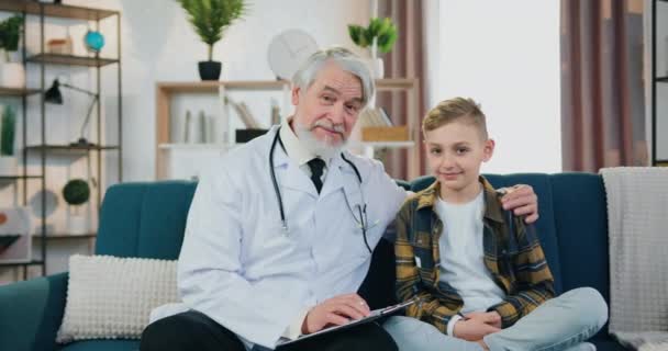 Detailní portrét dobře vypadající pozitivní respektované kvalifikované vousatý lékař a jeho dospívající chlapec pacient, který pózuje na kameře na pohodlné pohovce v útulné místnosti — Stock video