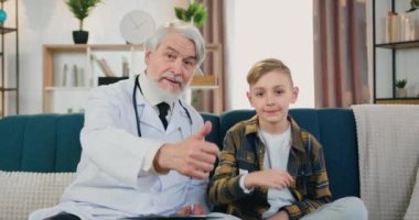 Çekici, nitelikli, sakallı, sakallı bir çocuk doktorunun gülümseyen genç bir çocukla kameraya baktığı ve baş parmak işareti yaptığı sağlık hizmeti konsepti.