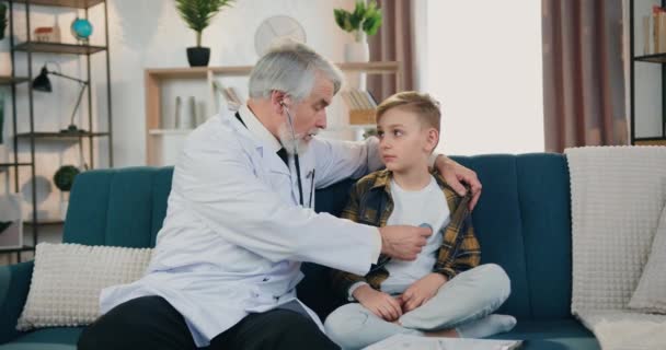 Aanzienlijke zelfverzekerde gekwalificeerde volwassen bebaarde arts gebruikt stethoscoop om hartslag en longen van jongen patiënt te onderzoeken tijdens huisbezoek, kind gezondheidszorg concept — Stockvideo