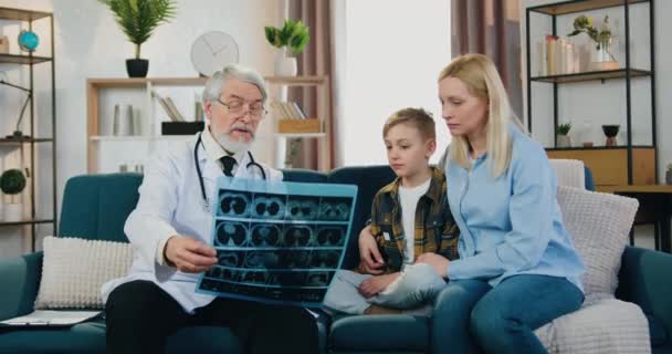 Krásná pozorná blond žena sedí na gauči spolu se svým nemocným dospívajícím synem a poslouchá vysvětlení výsledků chlapeckých rentgenových snímků od zkušeného respektovaného vousatého rodinného lékaře