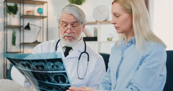Врачи дома посетить концепцию, где привлекательный уверенно опытный старший бородатый врач обсуждают результаты рентгеновского сканирования с красивой обеспокоенной пациенткой — стоковое видео