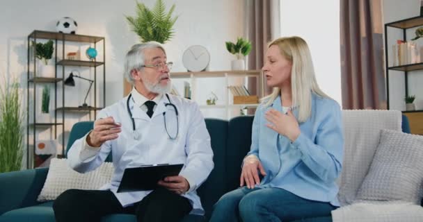 Egészségügyi koncepció, ahol jóképű figyelmes képzett érett szakállas orvos hallgat zaklatott szőke nő, amely azt mondja, egészségügyi panaszokat az ő otthoni látogatás — Stock videók