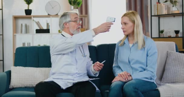 Pravděpodobný kvalifikovaný starší rodinný lékař kontrolující teplotu pacientek pomocí infračerveného bezkontaktního teploměru při domácí návštěvě, pohled zepředu — Stock video