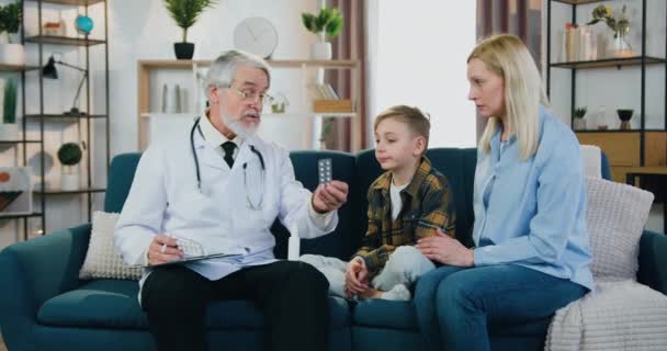 Привлекательный заботливый квалифицированный, уважаемый бородатый врач, дающий таблетки блондинке и ее сыну и объясняющий правила приема лекарств во время своего домашнего визита — стоковое видео