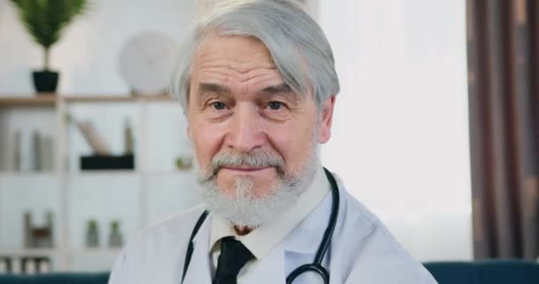 Close-up portret van knappe glimlachende succesvolle professionele gerespecteerde bebaarde arts in witte jas met stethoscoop die op zoek in de camera met zelfverzekerde gezichtsuitdrukking binnen — Stockvideo