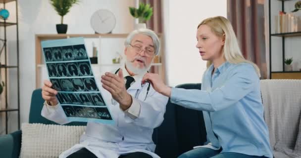 Médico barbudo respetado profesional responsable de buen aspecto que explica los resultados de la imagen de rayos X a la mujer que cuida bastante serio durante su visita a casa, de cerca — Vídeo de stock