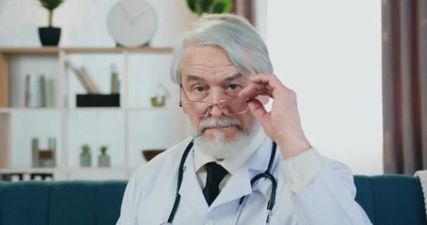 Närbild porträtt av sympatisk allvarlig kvalificerad framgångsrik senior skäggig läkare i glasögon som poserar på kameran på samtida hem interiör bakgrund — Stockvideo