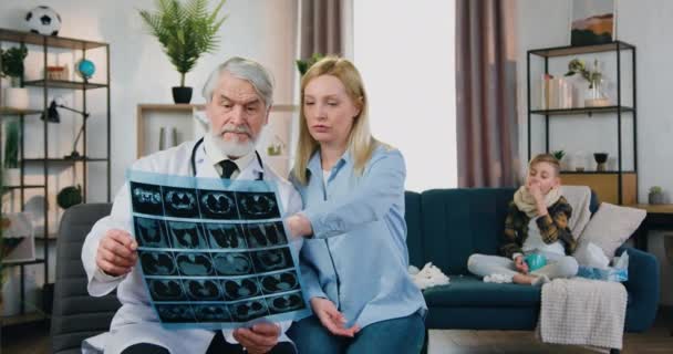 Roztomilý nemocný vyčerpaný chlapec s kašlem a bolestí v krku sedí na gauči, zatímco jeho pečující milující milující matka přezkoumává rentgen scan spolu se zkušeným starším vousatý lékař — Stock video