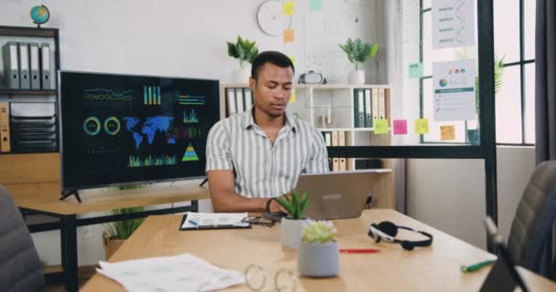 Beau prévenant qualifié intelligent afro-américain travaillant sur ordinateur portable assis sur son lieu de travail seul dans un bureau moderne le jour, vue de face — Video