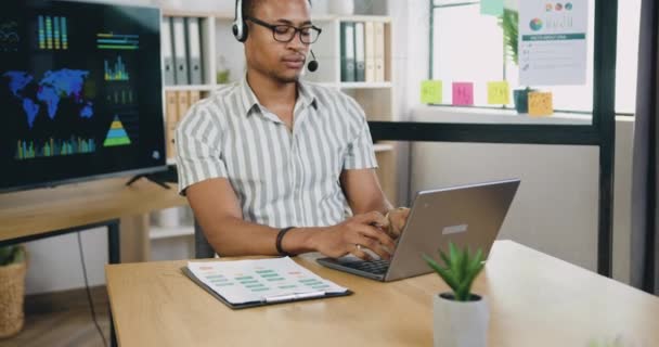 Atraente sério experiente bem sucedido jovem trabalhador de escritório afro-americano com fone de ouvido fazendo anotações em relatório financeiro do computador e olhando para a câmera no escritório — Vídeo de Stock