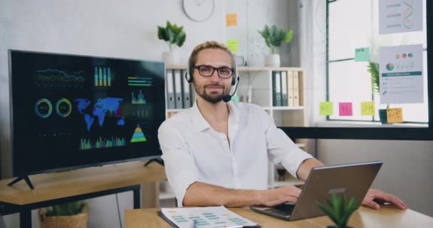 Portrét dobře vypadajícího usměvavého spokojeného úspěšného zkušeného kancelářského pracovníka v bezdrátové sluchátkové soupravě, který pózuje na kameře s palcem nahoru při práci na počítači — Stock video