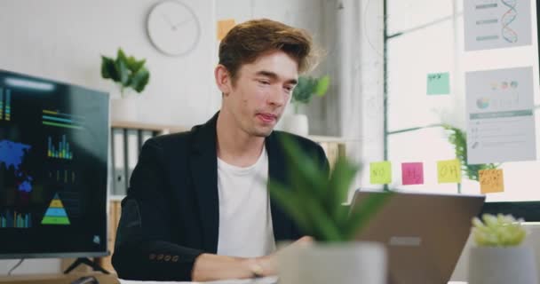 Närbild av snygg leende nöjd kreativ skicklig ung kille i casual kläder som arbetar på datorn i mysiga kontorsrum på dagtid — Stockvideo