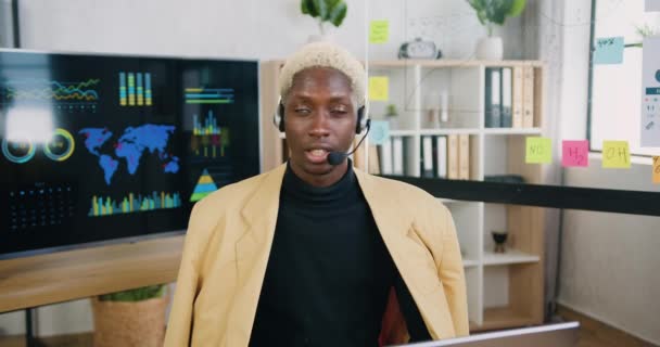 Sannolikt elegant säker erfaren afrikansk amerikansk manlig kontorsarbetare med hörlurar talar in i kameran till internet publik eller inspelning intervju på kontoret — Stockvideo