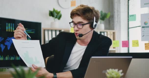 Atractivo positivo confiado experimentado joven trabajador de oficina con auriculares que explican informe con gráficos para sus colegas durante la videoconferencia en el ordenador portátil de la oficina — Vídeos de Stock