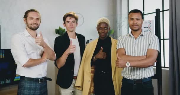 Attrayant sourire équipe masculine créative multiethnique réussie debout devant la caméra dans le bureau moderne et montrant pouces levés, vue de face, ralenti — Video