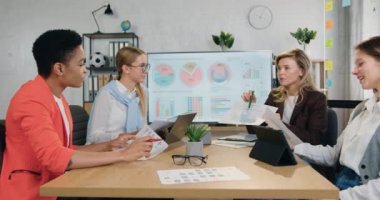 Çalışma masasında oturan genç iş kadınlarından oluşan bir ekip, programları ve yeni proje fikirlerini tartışıyor, şirket toplantısında deneyimleri ve yeni bilgileri paylaşıyor.