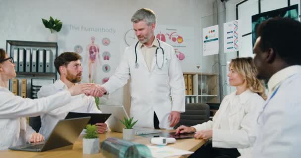 Arbetsflöde på läkarmottagningen där stilig säker kvalificerad skäggig läkare hälsar varje medlem av hans multietniskt team med handslag innan den gemensamma konferensen inleds — Stockvideo