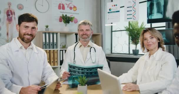 Retrato de boa aparência positiva bem sucedida experiente multiétnico masculino e feminino médicos que posando na câmera sentado em torno do local de trabalho no escritório da clínica — Vídeo de Stock