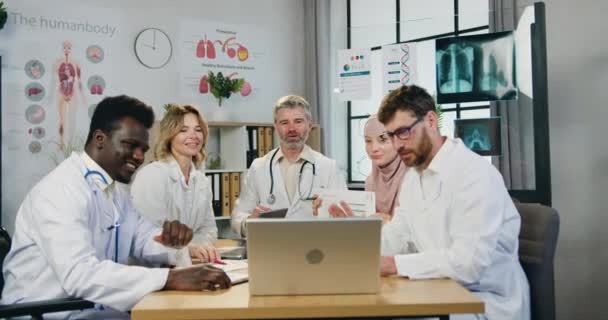 Jóképű pozitív sikeres tapasztalt, változatos férfi és női orvosok online eligazítás laptop a modern munkateremben, elölnézet — Stock videók