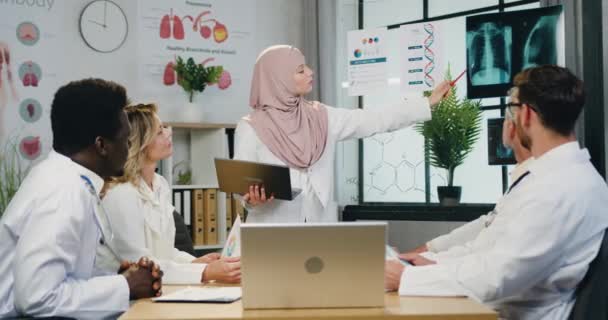 Atractivo médico arábiga cualificado seguro de sí mismo en hiyab explicando los resultados del diagnóstico por rayos X para los colegas masculinos y femeninos hábiles atantivos diversos durante el consejo conjunto — Vídeos de Stock