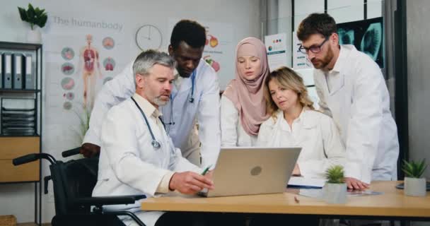 Beau succès respecté barbu médecin principal en fauteuil roulant travaillant avec ses collègues de travail multiethniques masculins et féminins confiants et hautement qualifiés en utilisant les informations sur l'ordinateur portable — Video