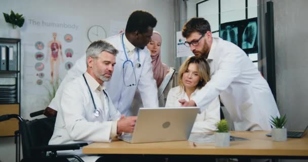 Ομάδα συμπαθής αυτοπεποίθηση υπεύθυνος έμπειρους διαφορετικούς γιατρούς που εργάζονται με πληροφορίες στην οθόνη του φορητού υπολογιστή, όταν θηλυκό αραβικό βοηθητικό πλησιάζει να γενειοφόρος κύριος γιατρός για να υπογράψει τα έγγραφα — Αρχείο Βίντεο