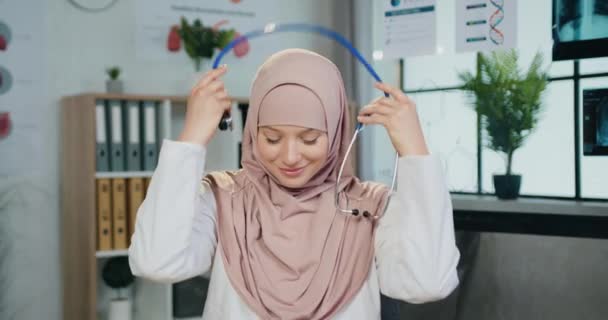 Portret van mooie tevreden succesvolle hooggeschoolde moslim vrouwelijke arts in hijab die stethoscoop op haar nek terwijl het kijken in de camera met oprechte glimlach in het ziekenhuis kantoor — Stockvideo