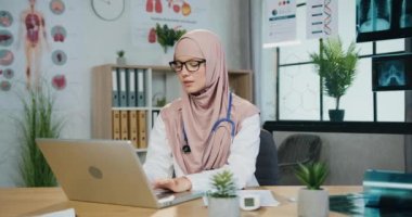 Çekici, dost canlısı, tesettürlü Müslüman kadın doktor klinikteki çalışma odasında bilgisayar üzerinde çalışıyor ve kameraya samimi bir gülümsemeyle bakıyor.