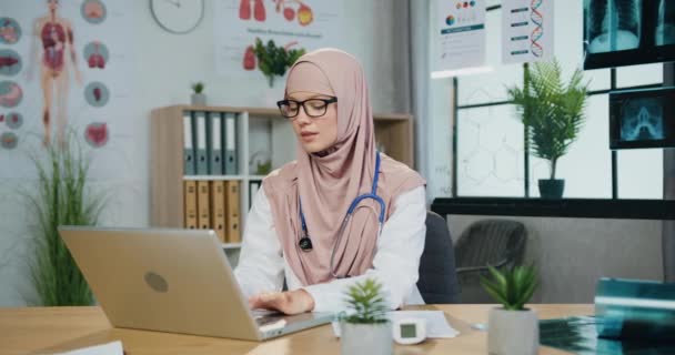 Atrakcyjny pozytywny i przyjazny wykwalifikowany muzułmanin lekarz w hidżabie pracuje na komputerze w swojej pracowni w klinice i patrząc w aparat z szczerym uśmiechem, zbliżenie — Wideo stockowe