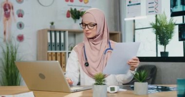 İyi görünümlü, kendine güveni yüksek, başörtüsü takan ve klinikte bilgisayarla ve raporlarla çalışan başarılı, kendine güvenen Arap kadın doktorların portresi.