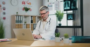 Güvenilir, güvenilir, deneyimli, sakallı, gri saçlı bir doktor. İş yerinde, sağlık dolabında oturuyor ve bilgisayar üzerinde çalışıyor.