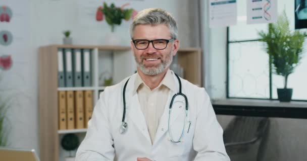 Portrét sympatického usměvavého přátelského úspěšného zkušeného vousatého lékaře v brýlích, který se dívá do kamery se spokojeným výrazem obličeje na interiéru kliniky — Stock video