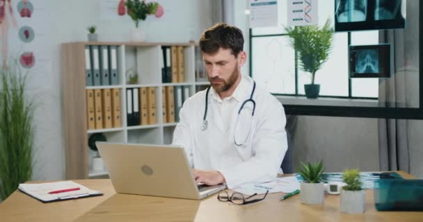 Flux de travail dans le bureau médical où beau sérieux confiance expérimenté médecin barbu tapant sur ordinateur, entrer les informations nécessaires dans le rapport médical — Video