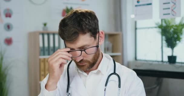 Dobře vypadající pozitivní sebevědomý kvalifikovaný vousatý lékař si sundává brýle a dívá se přitom do kamery s přívětivým výrazem ve tváři v moderní lékařské ordinaci — Stock video