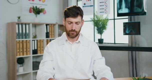 Atraktivní usměvavý úspěšný přátelský kvalifikovaný vousatý zdravotník v uniformě kliniky se stetoskopem pózování na kameře na nemocničním pracovišti pozadí — Stock video
