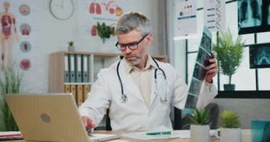 Güvenilir, sorumluluk sahibi, gözlüklü, sakallı bir doktor röntgen taramasının sonuçlarını bilgisayara giriyor, tıbbi çalışma odasında çalışıyor, kapatıyor.