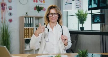 Güzel görünüşlü gülümseyen, başarılı, yetenekli, gözlüklü, sarışın, güzel dekore edilmiş bir muayenehanede kameraya poz veren ve baş parmağını kaldıran başarılı bir kadın doktor.