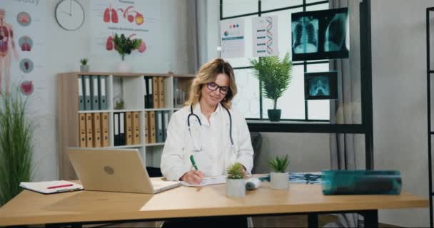 Krásná pozitivní sebevědomá zkušená blondýnka lékařka v brýlích a nemocniční uniformě pracující s papírovými zprávami v ordinaci, pracovní postupy na klinice — Stock video