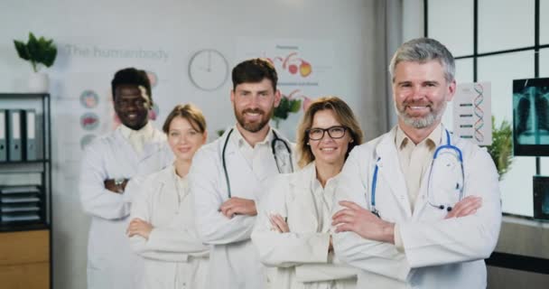 Aanzienlijke positieve professionele vriendelijk ervaren vijf mannelijke en vrouwelijke multi-etnische medische werkers poseren op camera met gekruiste armen in het ziekenhuis werkkamer, close-up — Stockvideo