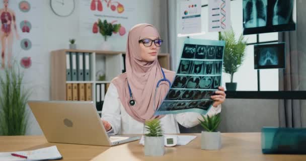 Medizinkonzept, bei dem eine ziemlich positive, selbstbewusste, erfahrene muslimische Ärztin im Hijab die Ergebnisse von Röntgenuntersuchungen und der Eingabe von Daten in den Computer in der Klinik lernt — Stockvideo