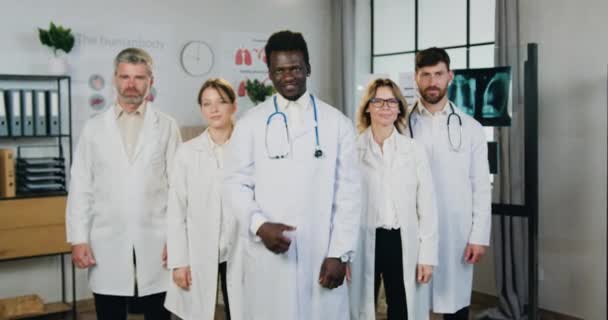 Likable uśmiechnięty pewny siebie profesjonalny zróżnicowany zespół lekarzy mężczyzn i kobiet stojących przed kamerą w gabinecie medycznym i pokazujących kciuki do góry, zbliżenie — Wideo stockowe