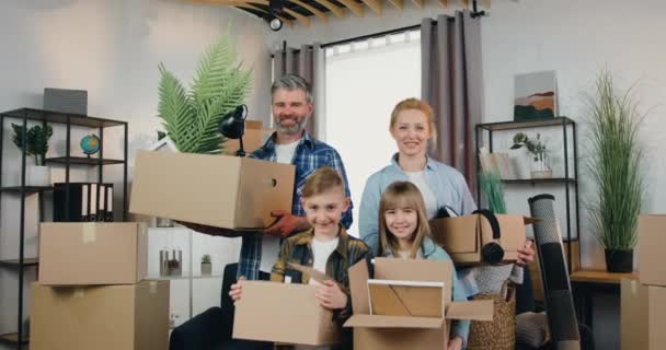 Áthelyezés koncepció, ahol szerethető mosolygós boldog modern család négy személy áll a kamera előtt kartondobozokkal az új lakásban — Stock videók