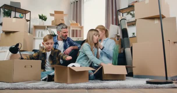 Agradable sonrisa feliz amigable familia moderna con dos niños sentados en el suelo entre cajas de cartón en el día de reubicación y desempacar diferentes decoraciones para el hogar — Vídeo de stock