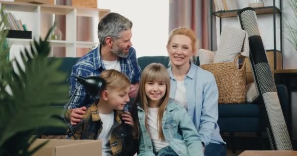 Heerlijk vrolijk gelukkig vriendelijk paar met twee kinderen poseren op camera met oprecht glimlachen op kartonnen dozen achtergrond als ze verhuizen naar een nieuw appartement, vooraanzicht — Stockvideo